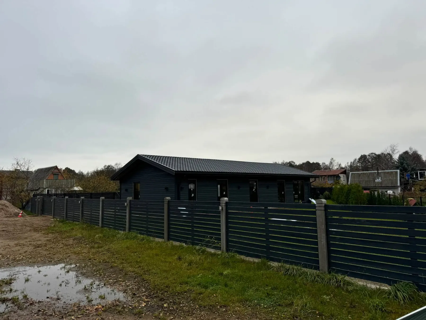 Pelēka vienstāva māja un horizontālu dēļu žogs ap zemesgabalu